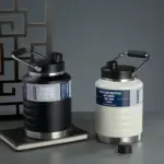 Dos termos para camping de 1500 ml de acero inoxidable con asa, uno negro sobre un cuaderno y otro blanco con la tapa abierta al lado.