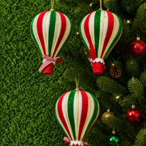 Tres adornos navideños colgantes con forma de globo aerostático, decorados con franjas verticales rojas, blancas y verdes, sobre un árbol de Navidad.