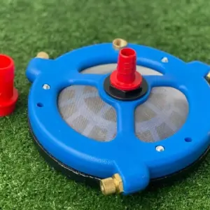 Mezclador y filtro agrícola azul con malla gris, conectores de latón y conector rojo sobre césped sintético.