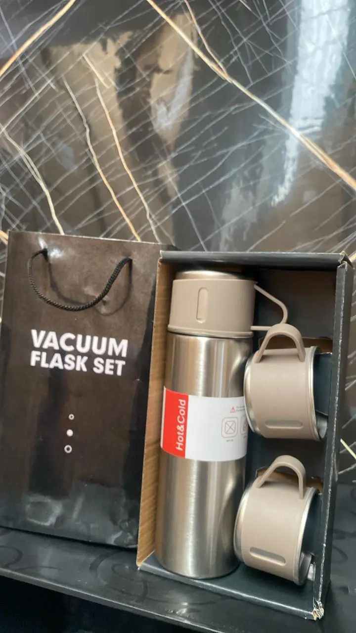 Juego de termo cilíndrico en acero inoxidable color plateado con dos tazas beige apiladas y bolsa de regalo negra con la frase vacuum flask set.