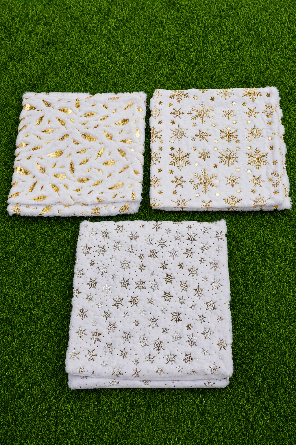 Tres cojines de pelo sintético blanco con estampados dorados de hojas y copos de nieve, sobre césped verde.