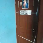 Forro textil para puerta color café con diseño del Club América, decorado con imágenes religiosas y asegurado con cintas blancas en una pared azul.