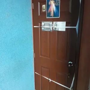 Forro textil para puerta color café con diseño del Club América, decorado con imágenes religiosas y asegurado con cintas blancas en una pared azul.