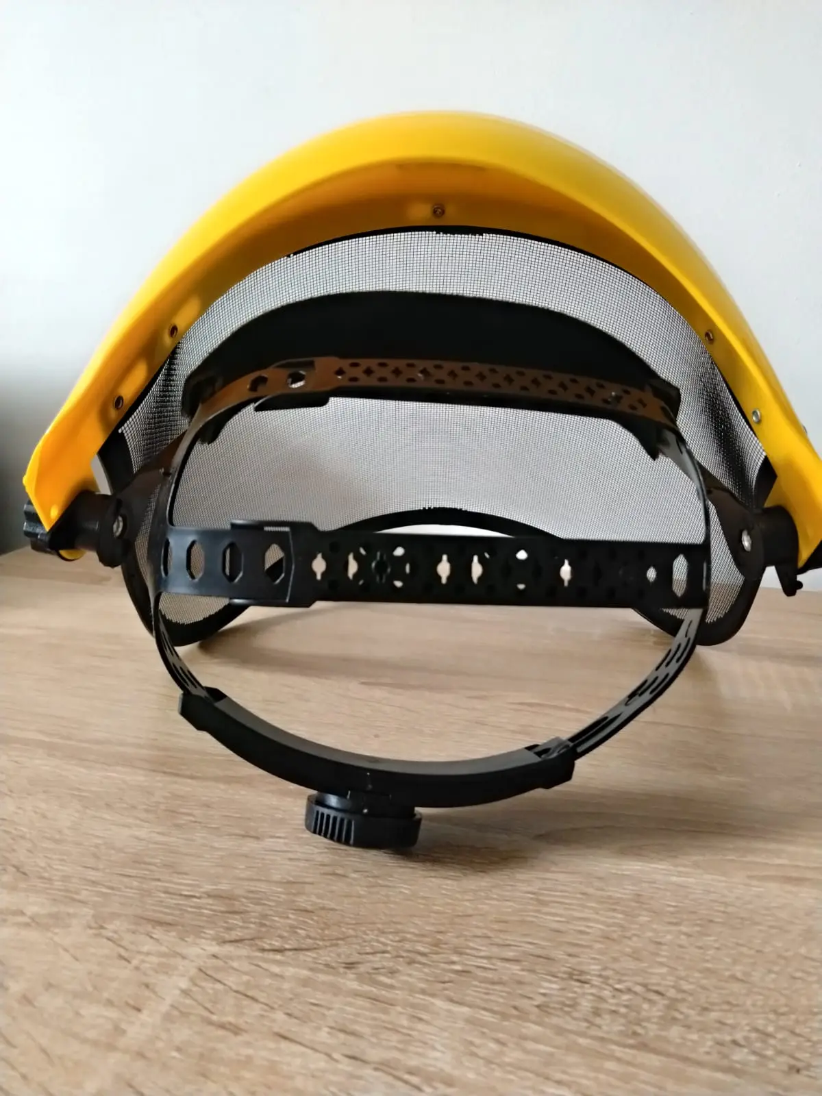 Careta de protección facial con malla metálica plateada, casco amarillo y arnés de ajuste negro para guadañadora.