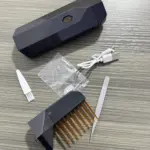 Peine eléctrico difusor de incienso negro con dientes dorados, estuche, cable USB, pinzas y espátula sobre superficie de madera gris.