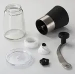 Despiece de molino de café manual negro con cuerpo de vidrio transparente, manivela metálica, muela cerámica blanca y piezas separadas sobre superficie blanca.