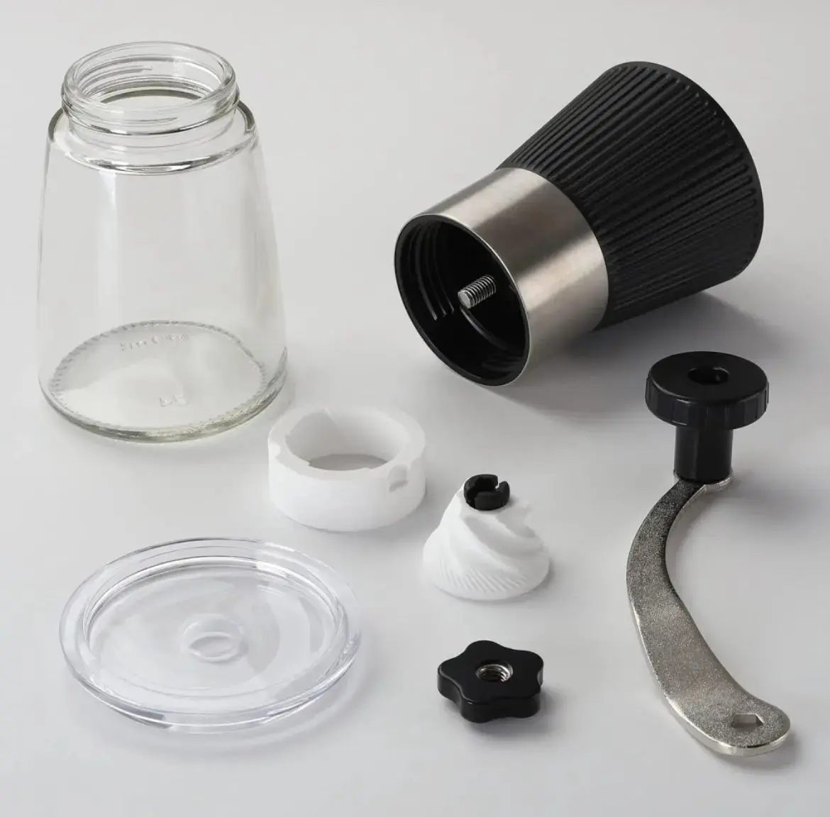 Despiece de molino de café manual negro con cuerpo de vidrio transparente, manivela metálica, muela cerámica blanca y piezas separadas sobre superficie blanca.