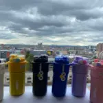 Seis termos de acero inoxidable de 520 ml en colores blanco, amarillo, negro, azul, morado y rosado, con tapa plástica transparente, alineados sobre una superficie blanca frente a una ventana con vista a la ciudad.