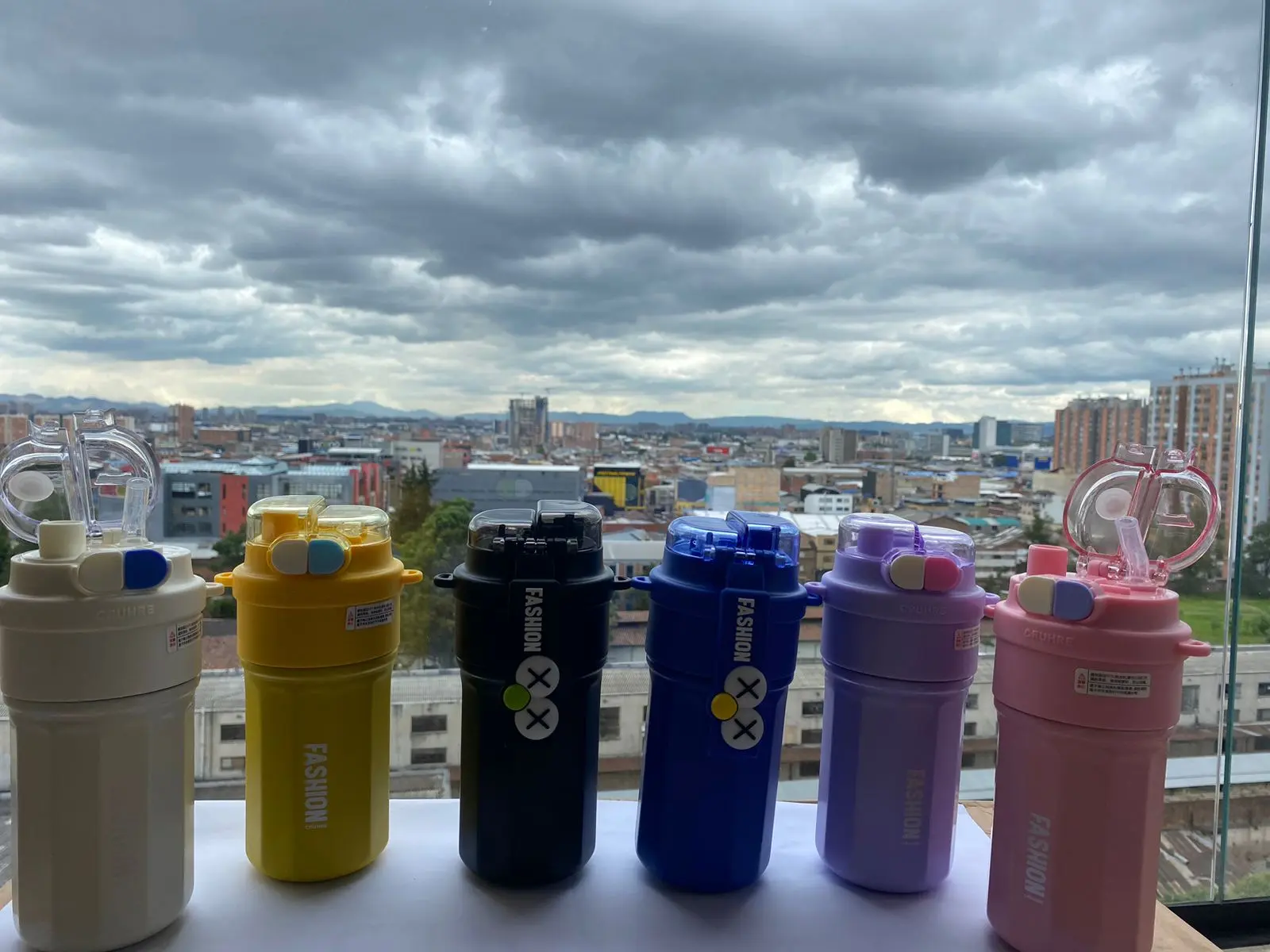 Seis termos de acero inoxidable de 520 ml en colores blanco, amarillo, negro, azul, morado y rosado, con tapa plástica transparente, alineados sobre una superficie blanca frente a una ventana con vista a la ciudad.