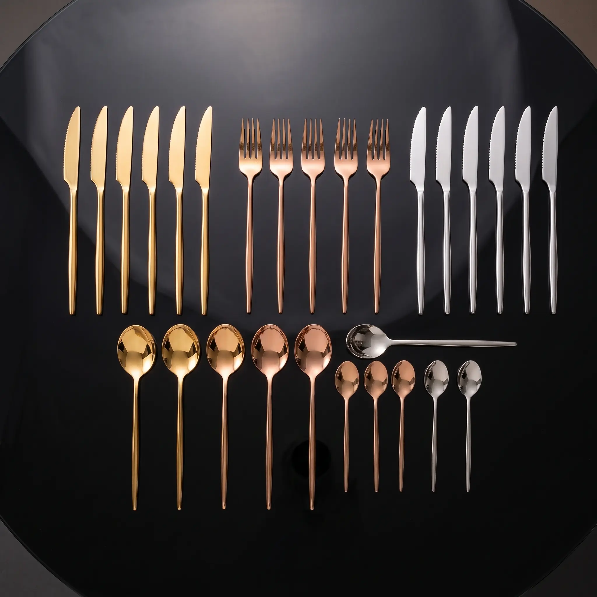 Juego de cubiertos de acero inoxidable con acabado brillante en tonos dorado, oro rosa y plateado, dispuestos ordenadamente sobre superficie oscura.