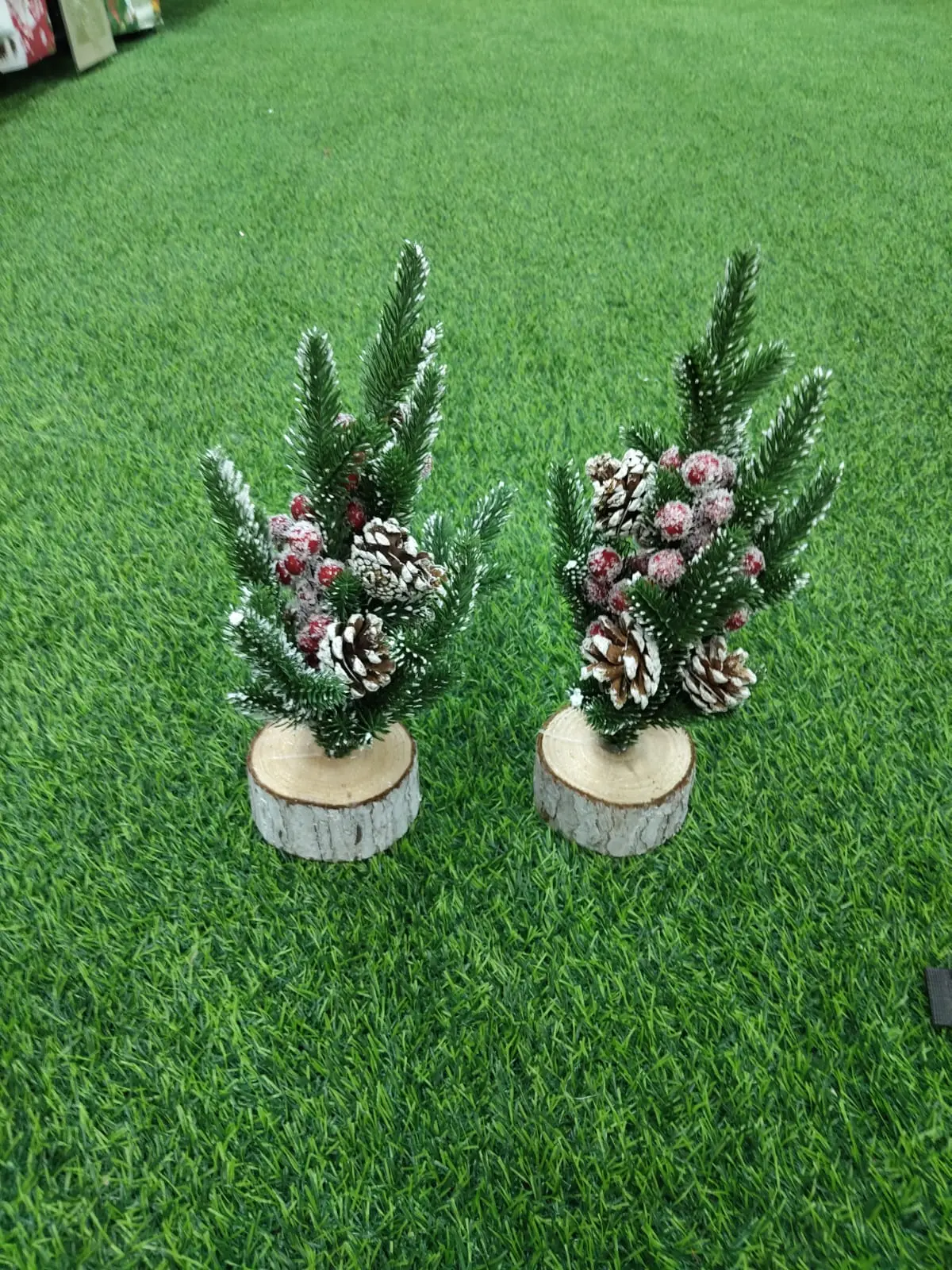 Dos mini pinos navideños artificiales con base de madera, decorados con piñas naturales y bayas rojas nevadas sobre césped sintético.