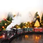 Tren eléctrico infantil con locomotora negra, vagones verde y rojo, efecto de humo blanco y luces cálidas en un decorado navideño.