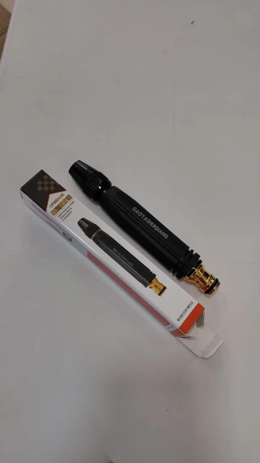 Boquilla pulverizadora ajustable de color negro con conector dorado, junto a su caja rectangular blanca y roja sobre superficie clara.