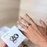 Mano con anillo inteligente plateado puesto y caja blanca con modelos en negro y plata para monitoreo biométrico.