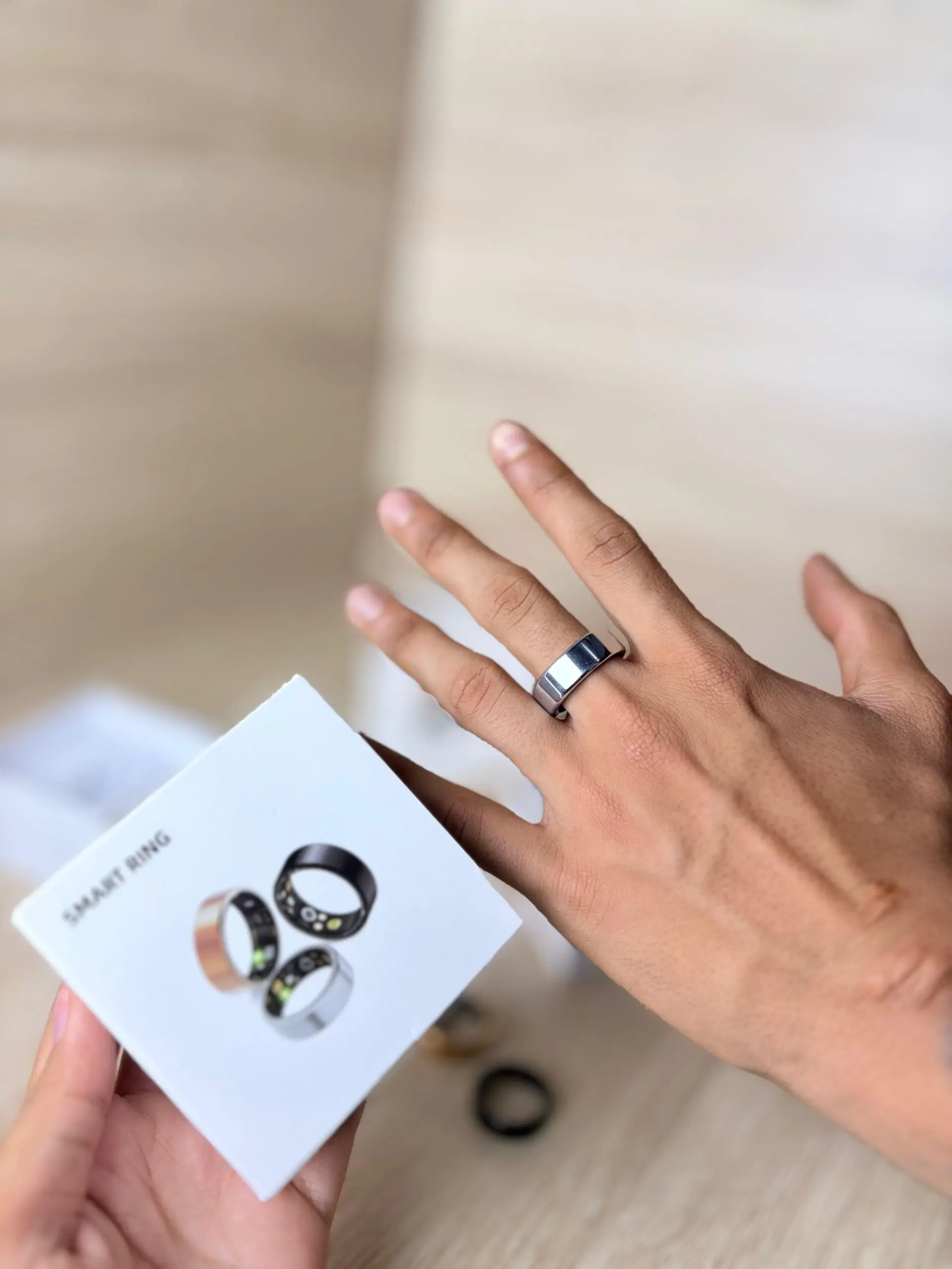 Mano con anillo inteligente plateado puesto y caja blanca con modelos en negro y plata para monitoreo biométrico.