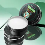 Tarrina negra abierta con mantequilla blanca para tatuajes Sadoer y tapa decorada con venado junto a base metálica.