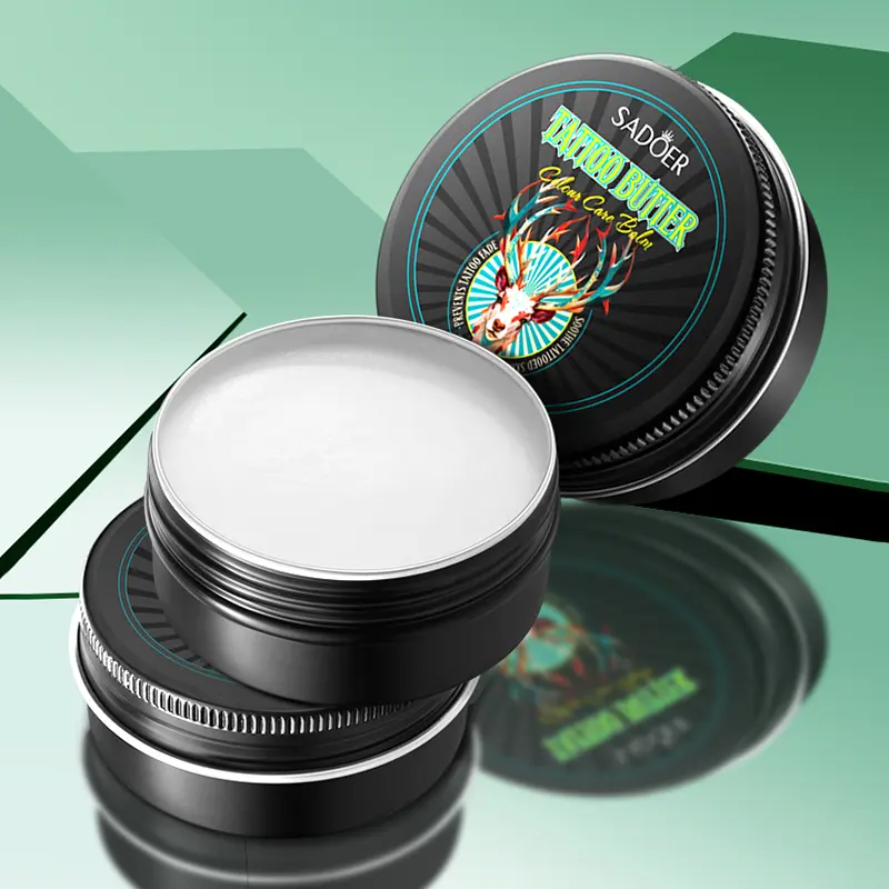 Tarrina negra abierta con mantequilla blanca para tatuajes Sadoer y tapa decorada con venado junto a base metálica.