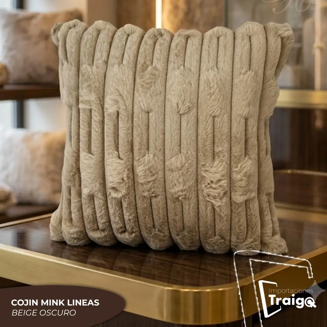 Cojín decorativo color beige oscuro con textura de peluche tipo mink y diseño de líneas verticales en relieve, exhibido sobre una mesa con borde dorado.