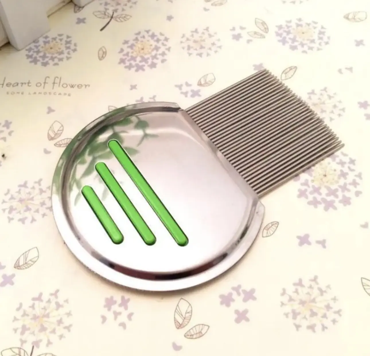 Peine antipiojos de acero inoxidable con mango circular plateado, tres líneas diagonales verdes y dientes metálicos finos y largos.