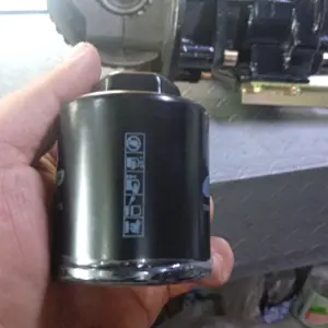 Filtro de aceite cilíndrico negro para motocarro AKT sostenido por una mano, con piezas mecánicas de motor al fondo.