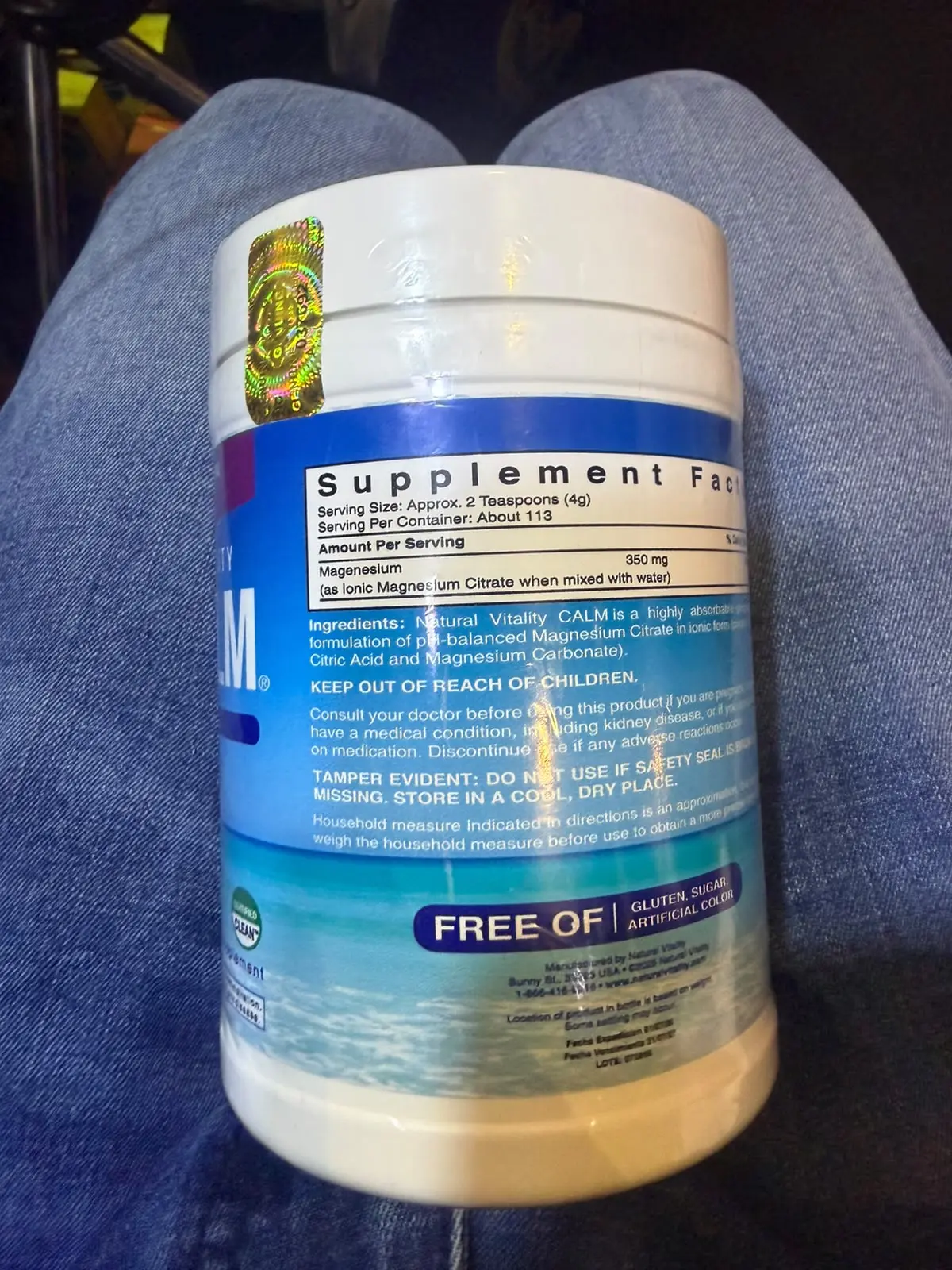 Envase blanco de suplemento de magnesio con etiqueta azul y blanca que muestra la tabla nutricional, sostenido sobre unos jeans azules.