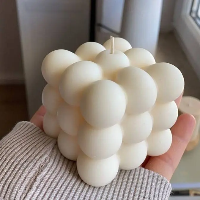 Vela aromática bubble de cera color marfil con diseño geométrico cuadrado, reposando sobre una mano en un espacio interior.