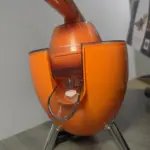Exprimidor eléctrico de naranjas color naranja con palanca superior, boquilla transparente y base de tres patas metálicas sobre mesa de madera.