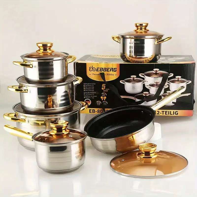 Batería de cocina en acero inoxidable brillante con detalles y asas doradas, incluye cacerolas con tapa de vidrio y sartén antiadherente organizadas junto a su caja.