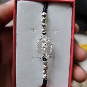 Pulsera ajustable de La Santa Muerte en hilo negro con abalorios plateados y dije ovalado rodeado de circonias, presentada en caja roja.