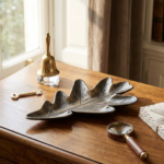 Bandeja decorativa de metal con acabado bronce envejecido en forma de hoja de roble, sobre escritorio de madera junto a una campana, llave y lupa.