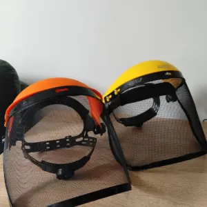 Set de dos caretas de malla para guadañadora con cabezales naranja y amarillo, visores negros y banda de ajuste, sobre superficie de madera.