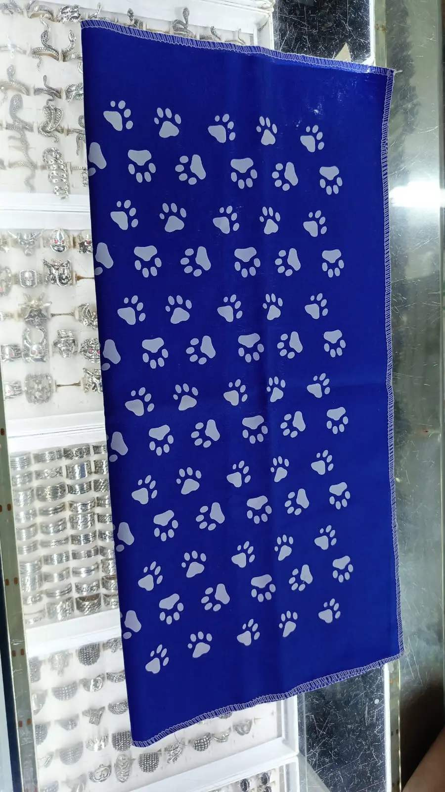 Pañoleta azul para perros con estampado repetido de huellas blancas, bordes cosidos y fondo de vitrina con joyería.