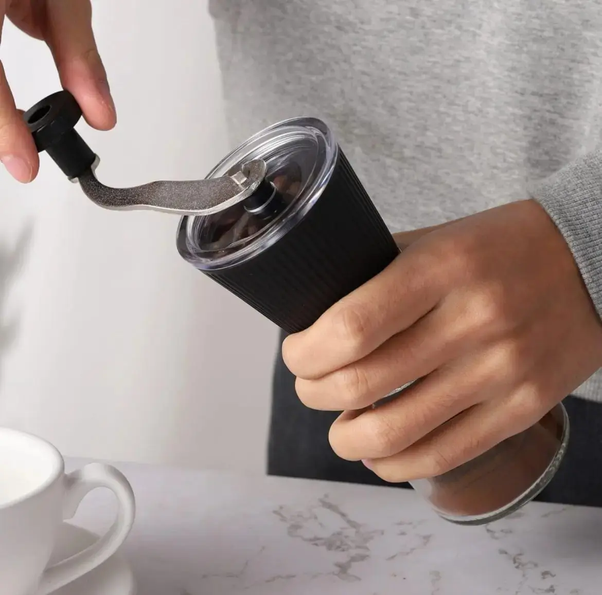 Molino de café manual negro con cuerpo de vidrio y muela cerámica, siendo accionado con manivela superior sobre una mesa.