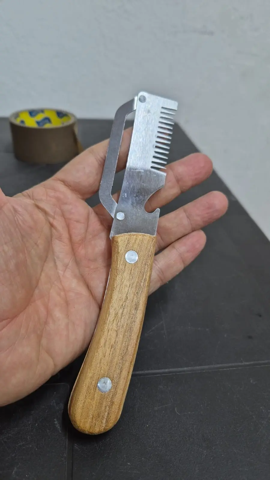 Pelador de verduras dentado con mango de madera, hoja de acero inoxidable en forma de peine y protector abatible, sostenido en una mano.