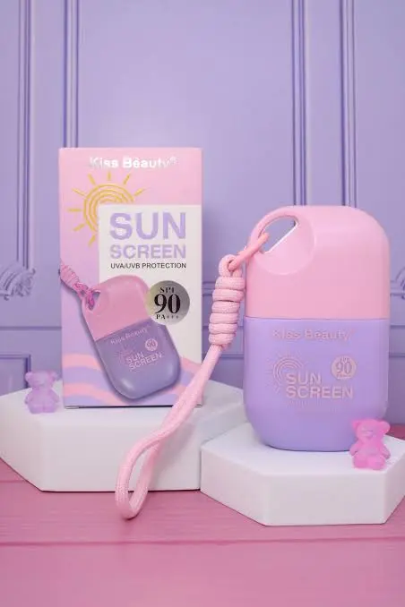 Protector solar facial Kiss Beauty SPF 90 portátil en tonos rosa y lila, con empaque, caja y cordón decorativo sobre soporte blanco.
