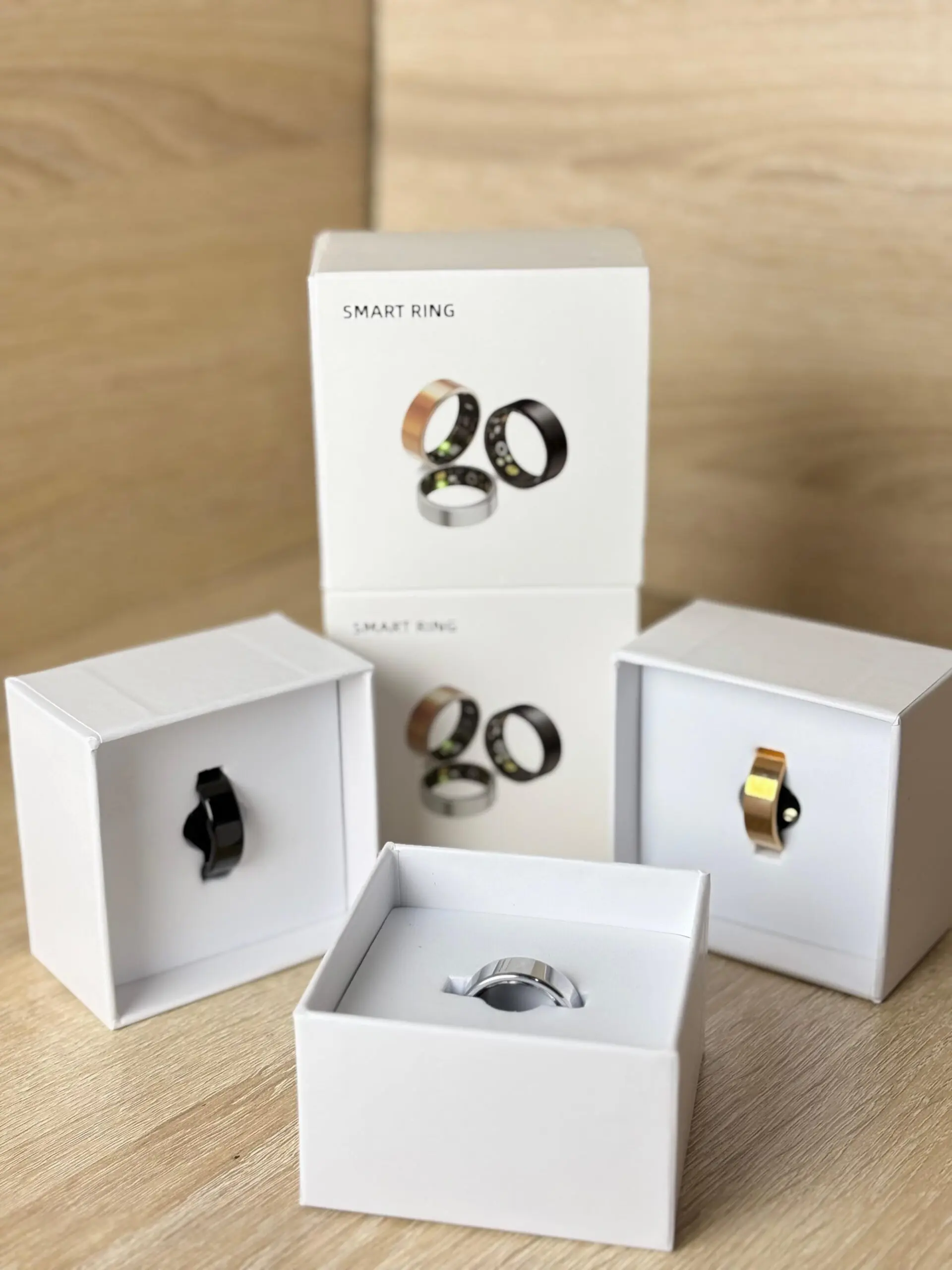 Anillos inteligentes de salud y monitoreo biométrico en cajas blancas, mostrando variantes en negro, dorado y plateado sobre fondo de madera.