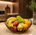 Frutera de hierro negro con bordes arqueados y base de madera, llena de frutas frescas sobre un mesón de cocina moderno.