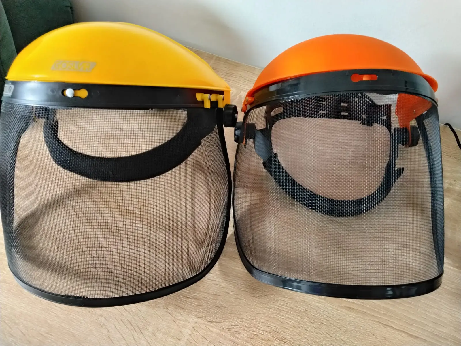 Set de dos caretas de malla para guadañadora con visores negros y cabezales de colores amarillo y naranja sobre superficie de madera.