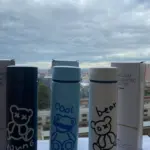Tres termos de acero inoxidable de 500 ml con diseños de osos y textos, colores negro, azul y blanco, frente a sus cajas y vista a la ciudad.