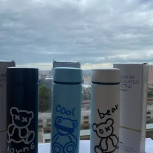 Tres termos de acero inoxidable de 500 ml con diseños de osos y textos, colores negro, azul y blanco, frente a sus cajas y vista a la ciudad.