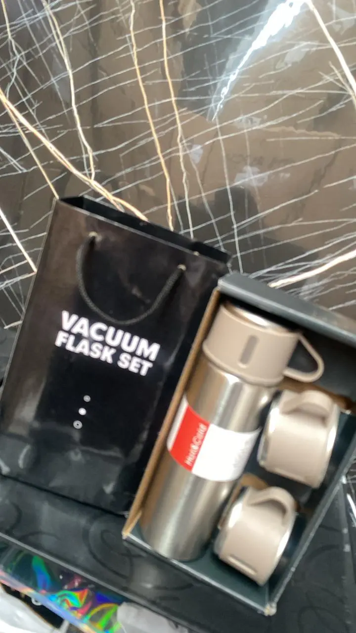 Juego de termo cilíndrico en acero inoxidable color beige y plata con dos tazas con asa, dentro de su caja junto a bolsa de regalo negra.