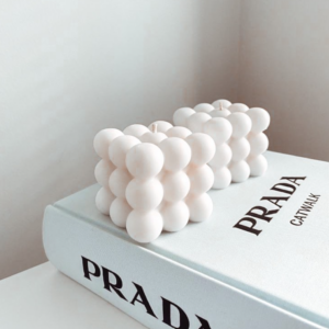 Par de velas blancas de cera con diseño geométrico de burbujas, sobre un libro blanco en un ambiente decorativo minimalista.