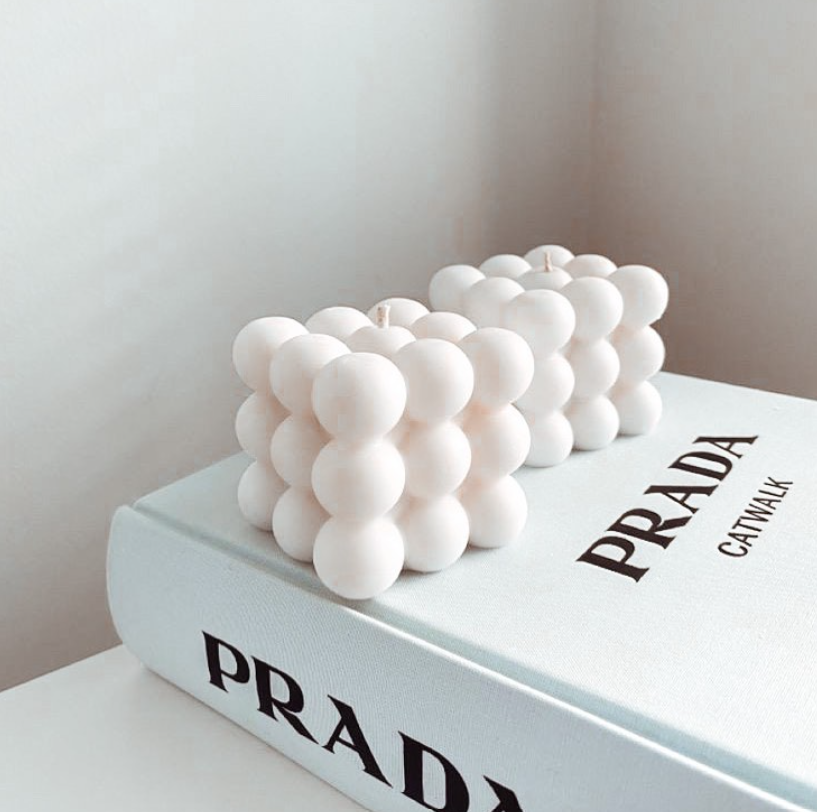 Par de velas blancas de cera con diseño geométrico de burbujas, sobre un libro blanco en un ambiente decorativo minimalista.
