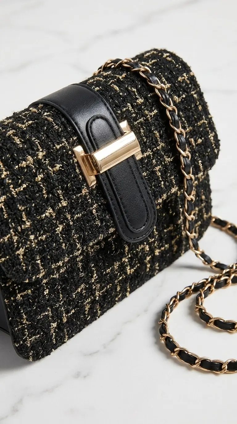 Bolso de tweed negro y dorado con solapa de cuero, cierre metálico dorado y correa de cadena entrelazada, visto en primer plano.