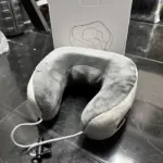Cojín de viaje ergonómico de tela gris aterciopelada con cable USB y conector, junto a su caja blanca sobre un piso oscuro.