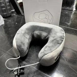 Cojín de viaje ergonómico de tela gris aterciopelada con cable USB y conector, junto a su caja blanca sobre un piso oscuro.