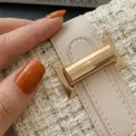 Bolso de mano en tweed crema con hilos dorados, solapa beige y hebilla metálica dorada en primer plano.