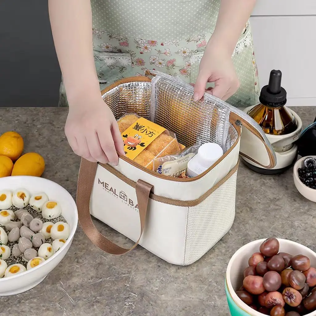 Maleta lonchera térmica color crema con asas café y revestimiento plateado, siendo llenada con pan y recipientes en una mesa de cocina.
